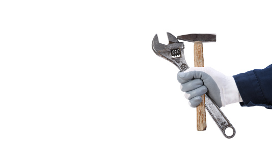 A worker's hand in a protective glove holds a large old adjustable wrench and a hammer on a white background.