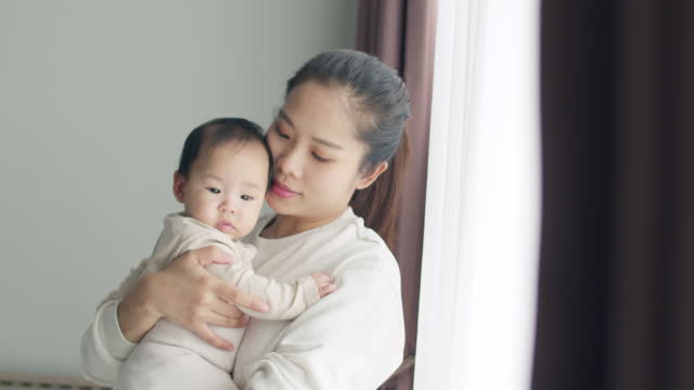 Asian mother holding a newborn baby in her arms beside the window at home.