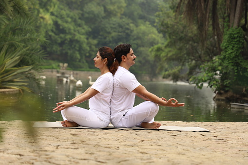 A beautiful young couple exercising and doing Yoga poses in a green serene environment early morning in a park to maintain a healthy lifestyle.