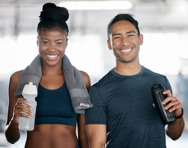 portrait, fitness et eau potable avec un couple de sportifs dans la salle de gym ensemble après une séance d’entraînement. diversité, sourire et serviette avec un jeune homme et une jeune femme heureux dans un centre de santé pour l’exercice ou l� - photography color image people real people photos et images de collection