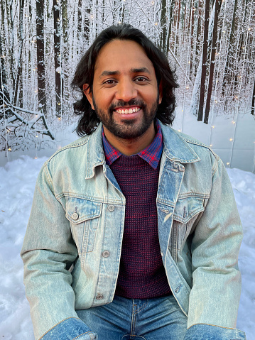 Stock photo showing portrait of Indian man casual clothing of denim jacket and jeans with jumper over collared against a background of seasonal winter display scene with artificial  snow.