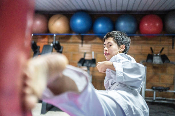 un garçon enfant donnant un coup de pied dans un sac de boxe pendant un cours de karaté - karate punching bag caucasian leisure activity photos et images de collection
