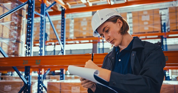 Young adult Caucasian woman warehouse worker working in factory warehouse, check merchandise package, writing clipboard. Logistic industry business, industrial job career, people at work concept