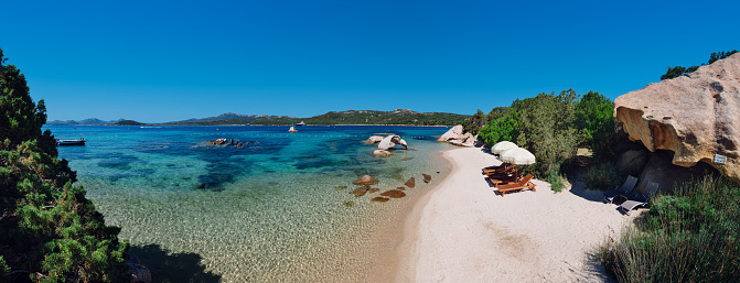 A stunning view of La Spiaggia dell'Elefante, a luxurious beach located in the Sardinia region of Sardinia. The wide-angle shot showcases the beach's crystal-clear, turquoise water that glistens in the bright summer sun. The fine white sand stretches out for miles, inviting visitors to take a leisurely stroll or relax under the warm Mediterranean rays or take a nap on the wooden lounge chairs.\n\nWe can see a cluster of rocky formations, one of which resembles an elephant. The natural stone formation adds a unique touch to the already picturesque landscape, making La Spiaggia dell'Elefante one of the most distinctive and memorable beaches in the world.\n\nThe overall scenery is breathtaking, with a perfect blend of natural beauty and human luxury. Visitors can indulge in various beach activities, such as swimming, sunbathing, snorkeling, or even taking a yacht out for a spin. La Spiaggia dell'Elefante is undoubtedly a must-visit destination for anyone looking for an unforgettable beach experience in the Mediterranean.