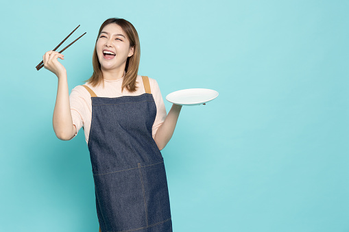 Young Asian woman housewife wearing kitchen apron cooking and holding empty white plate and chopsticks isolated on green background