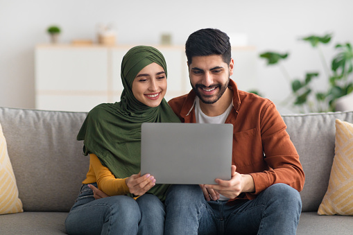 Happy Middle Eastern Spouses Using Laptop Watching Movie Online Or Browsing Internet Together Sitting On Couch At Home. Technology And Internet. Modern Muslim Family Lifestyle. Front View