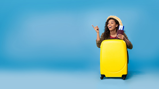 Carefree young black woman holding passport and tickets, sitting behind yellow suitcase, ready to travel on blue studio background, banner design with copy space. Summer vacation concept