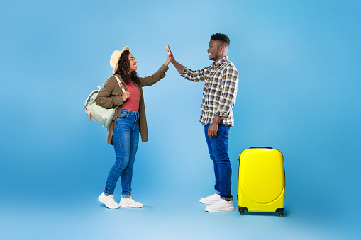 Full length of cool young black couple with luggage high fiving each other on blue studio background. Cheerful African American travelers with baggage going on summer vacation