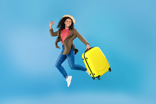 Excited young black woman in casual wear with bright suitcase jumping on blue studio background, copy space. Overjoyed African American female traveler with baggage flying up in air