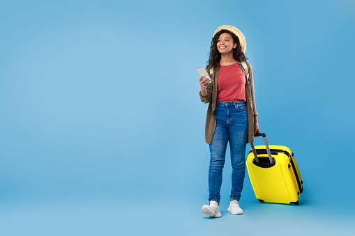 Young black female traveler with suitcase holding smartphone, booking hotel or vacation online, using internet travel agency app on blue studio background, empty space