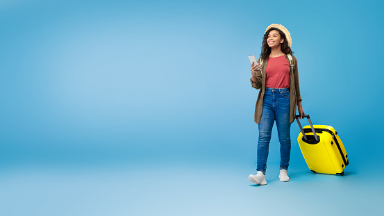 Cheery young black female traveler with suitcase holding smartphone, booking hotel or vacation online on blue studio background, banner design with empty space. Tourism, summer holidays concept