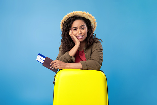 Upset young black woman holding passport and plane tickets, leaning on suitcase, missing vacation because of coronavirus lockdown on blue studio background. Unhappy lady being late for her flight