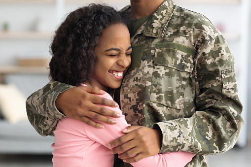 Closeup of happy black girl teenager hugging her mother soldier, smiling with closed eyes, kid enjoying reunion with mom, unrecognizable black woman in camouflage uniform returned home from army