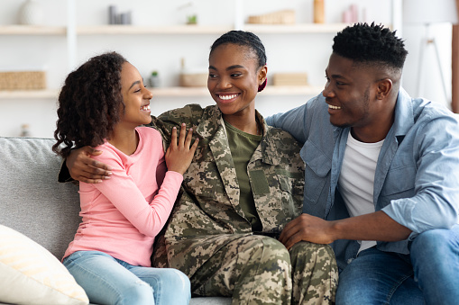 Loving african american family celebrating reunion with mother soldier, happy black girl teenager, woman in camouflage uniform and man sitting on couch at home, cuddling and having conversation