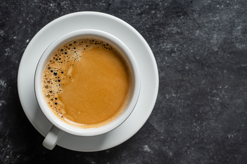 White porcelain coffee cup with saucer over black background, top view, copy space, close up. Hot coffee in a breakfast