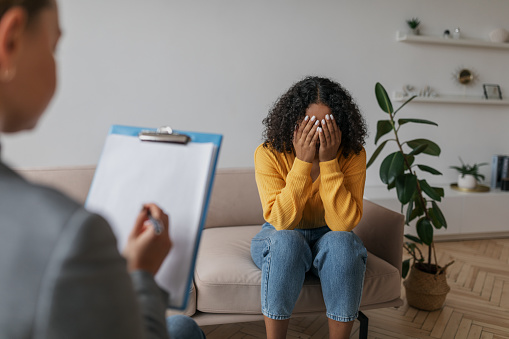 Unhappy young African American woman covering face with hands, crying on session with psychologist at office. Depressed black female speaking to mental health professional, having nervous breakdown