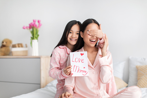 Satisfied chinese teenage girl closes eyes to young lady gives card and congratulates with mother day, sits on bed in bedroom interior. Surprise, holiday, love and relationship. Celebrate at home