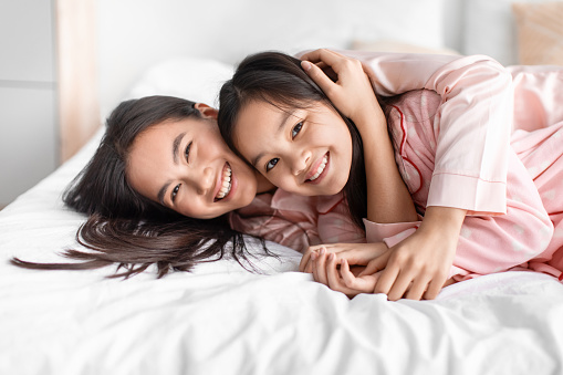 Glad happy pretty cute japanese young woman hug teen girl in pink pajama lie on bed and have fun in bedroom interior, free space. Love and relationship, mother and daughter together at home, close up
