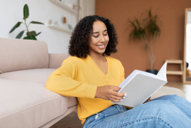 istock Smiling young black woman reading notebook, sitting on floor, getting ready for exam or business meeting at home 1480036388