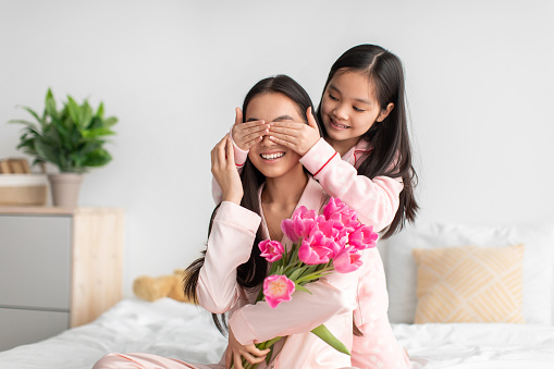 Smiling chinese adolescent girl closes eyes to millennial female with bouquet of tulips in bedroom interior. Congratulates mom on birthday, mother day at home, surprise for holiday at spring, indoor