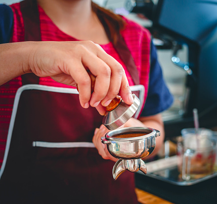 Hand of a barista holding a portafilter and a coffee tamper making an espresso coffee. Barista presses ground coffee using a tamper in a coffee shop