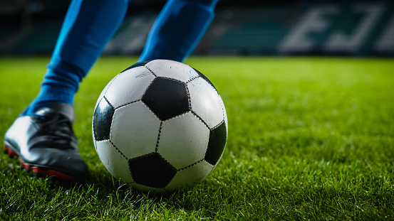 Close-up of a Leg in a Boot Kicking Football Ball. Professional Soccer Player Hits Ball with Fierce Power, Scores Goal, Grass Flying. Beautiful Cinematic Low Angle Ground Artistic Shot