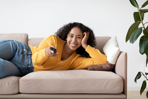 Young black woman relaxing on sofa, watching TV, switching channels with remote control, enjoying movie at home