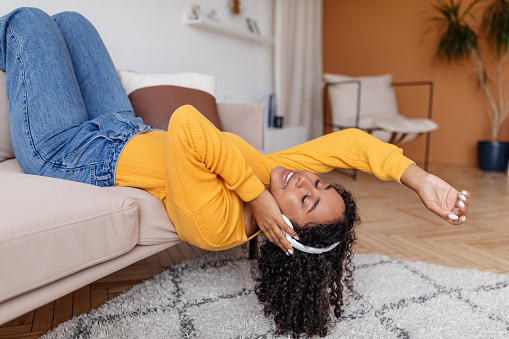 Cheerful young black woman in headphones lying on sofa with closed eyes, listening to music at home. Millennial African American female enjoying new audio book in headset, relaxing in living room