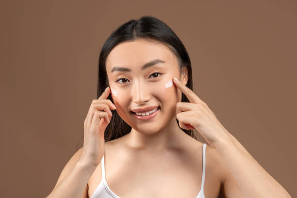 istock Attractive young asian lady touching her smooth cheeks, applying moisturizer cream and smiling at camera 1480040095