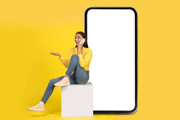 Excited Woman Talking On Cellphone Sitting Near Huge Smartphone, Studio Excited Woman Talking On Cellphone Sitting On Cube Near Huge Smartphone With Blank Screen Over Yellow Studio Background. Mobile Communication. Mockup, Collage giant cube stock pictures, royalty-free photos & images