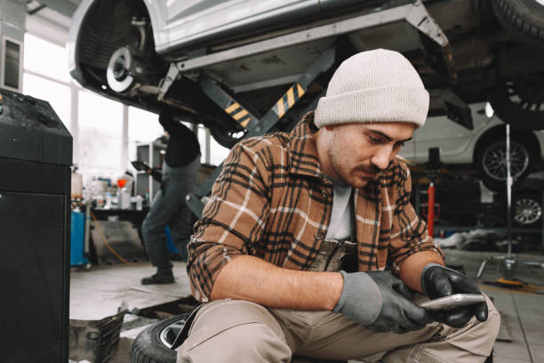 istock Car service worker sitting on a wheel and resting, using mobile phone 1480044802