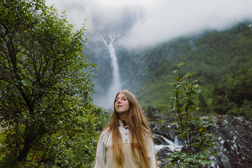 Low-Angle view of a woman explorer with long hair walking to dramatic Mardalsfossen waterfall hidden in the beautiful green mountain valley in Scandinavia