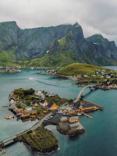 Drone high-angle photo of Sakrisoy village with yellow rorbu huts on the islands and bridge between them, surrounded by turquoise coloured sea and green mountain peaks on Lofoten, Nordland, Scandinavia