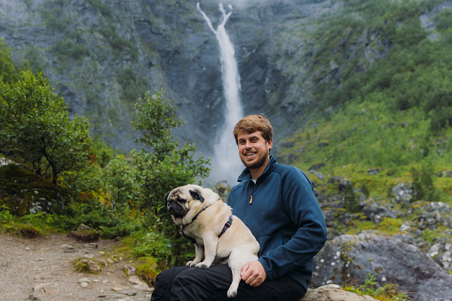 Happy male sitting with his pug with background view of dramatic Mardalsfossen waterfall hidden in the summer green valley in Scandinavia