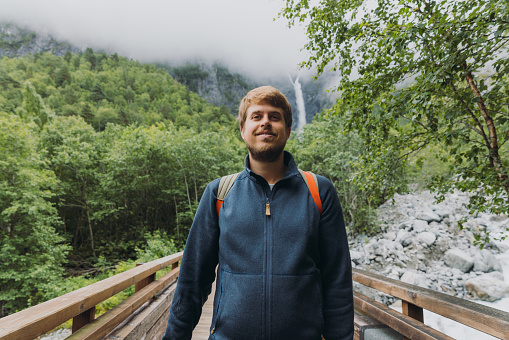 Happy male backpacker walking to dramatic Mardalsfossen waterfall hidden in the summer green valley in Scandinavia