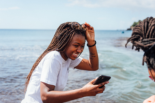Woman smiling and raising her sunglasses, with her phone in her hand, while at a beach