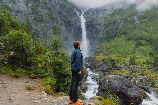 A Male backpacker walking to dramatic Mardalsfossen waterfall hidden in the summer green valley in Scandinavia