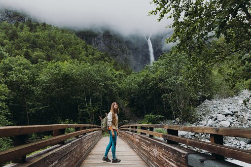 Happy woman backpacker walking to dramatic Mardalsfossen waterfall hidden in the summer green valley in Scandinavia