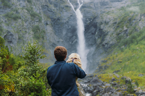 A male holding his pig enjoying a view of dramatic Mardalsfossen waterfall hidden in the summer green valley in Scandinavia