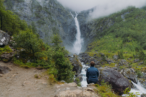 A male backpacker walking to dramatic Mardalsfossen waterfall hidden in the summer green valley in Scandinavia