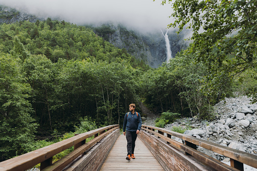 Happy male backpacker walking to dramatic Mardalsfossen waterfall hidden in the summer green valley in Scandinavia