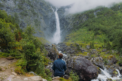A male backpacker walking to dramatic Mardalsfossen waterfall hidden in the summer green valley in Scandinavia