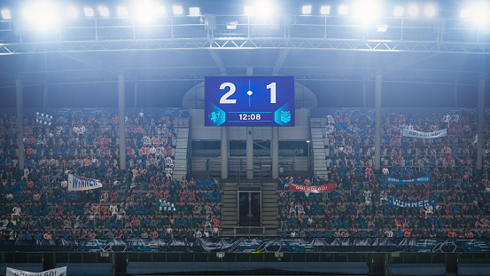 Football Soccer Stadium Championship Match, Scoreboard Screen Showing Score of 2:1. Crowd of Fans Cheering, Screaming, Having Fun. Sport Channel Television Advertising Concept. Wide Shot.