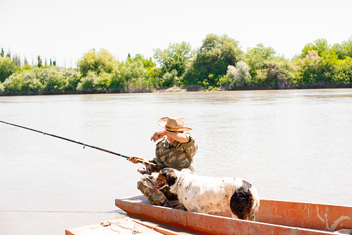 Tired fisherman in hat wiping sweat from forehead, while sitting in boat with dog and catching fish. Front view of exhausted man with fishing rod angling on river in hot day. Concept of fatigue.