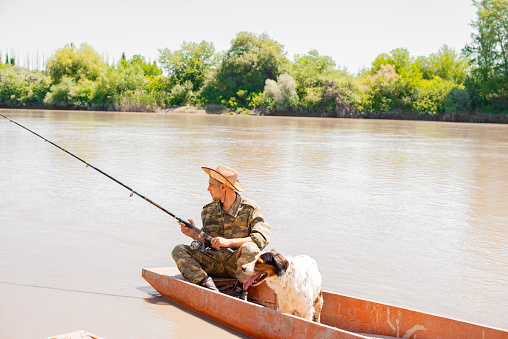 Relaxed angler in camouflage holding fishing pole, resting, while floating on lake with dog on board. Side view of pensive male fishing from boat, with landscape on background. Concept of fishery.