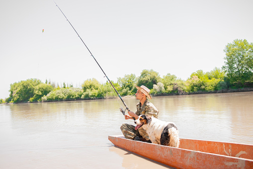 Professional fisherman checking hook by pulling rod out of water, while floating in old boat along river. Side view of young guy in camo fishing with dog, while resting on vacation. Concept of hobby.