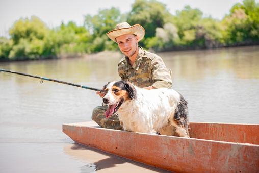 Smiling man in camouflage and hat fishing, while wading in muddy river on old boat. Front view of cheerful fishing with rod floating with spotted setter dog on board in sunny day. Concept of fishing.