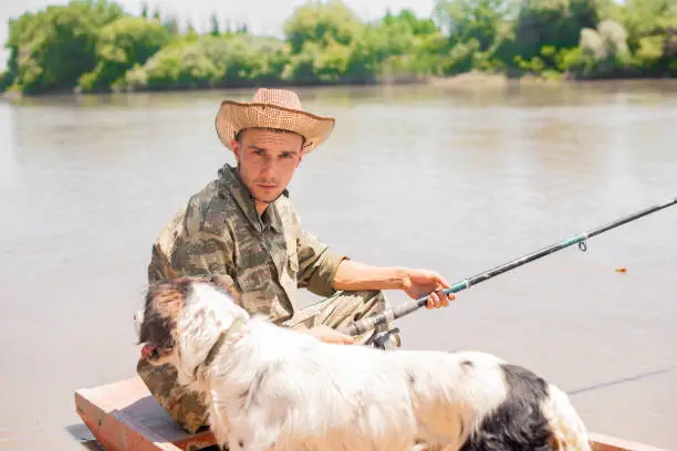 Photo of Portrait of young, serious fisherman with favourite dog, during calm fishery time.