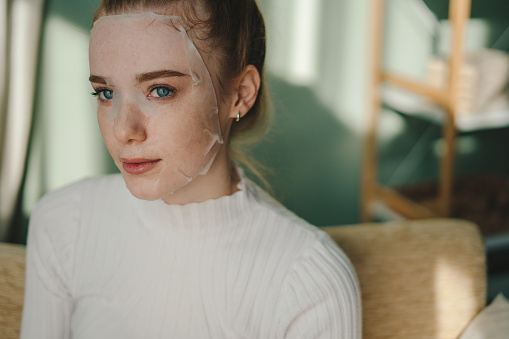 Portrait of beautiful caucasian ginger woman with paper sheet mask on her face looking contemplatively somewhere far away. Woman face skin care cosmetic. Medical healthcare. Young woman face portrait.