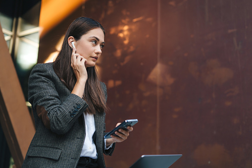 Brunette caucasian businesswoman putting earbud into her right ear sitting outdoors. Internet technology. Online business.